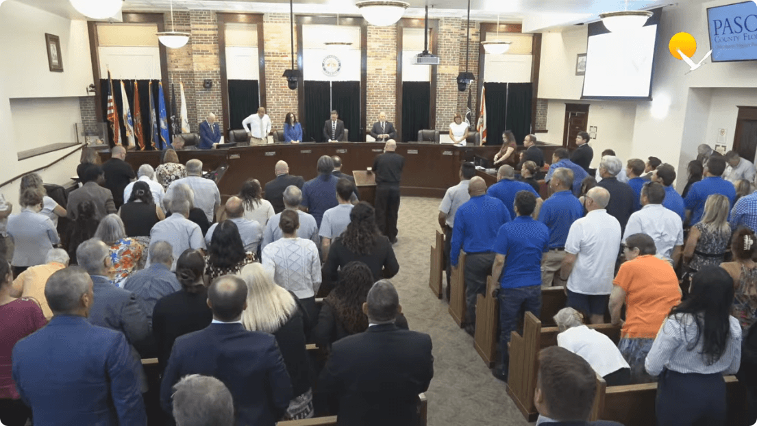 A packed government meeting chamber shows commissioners seated at a dais facing a standing crowd of attendees during a public session. Frame from the official Pasco County Government meeting recording at 0:00:34.