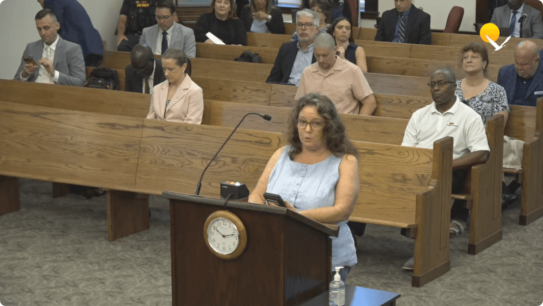 A woman speaks at a wooden podium in a public meeting chamber with an audience seated in gallery pews behind her. Frame from the official Pasco County Government meeting recording at 0:09:18.