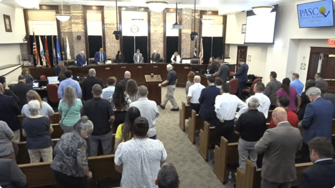 A packed government meeting room with commissioners seated at a dais and attendees standing, with a Pasco County Florida sign visible on screen. Frame from the official Pasco County Government meeting recording at 0:00:15.