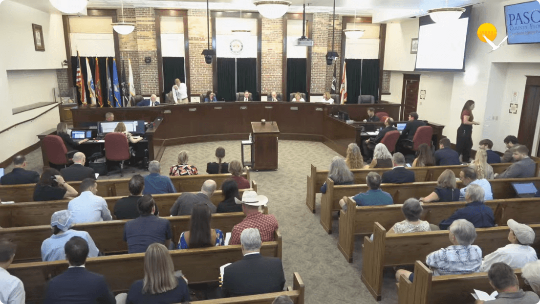 Wide shot of a government meeting room with commissioners seated at a dais and an audience filling wooden pews, watched by a Frame from the official Pasco County Government meeting recording at 0:00:16.